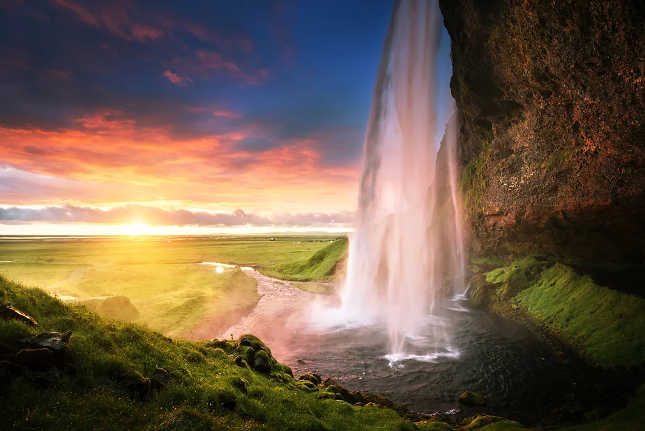 Plano general de una cascada islandesa durante un atardecer que baña el cielo de ese país de colores morados y rosados mientras los rayos de sol interceden en la caída del agua.