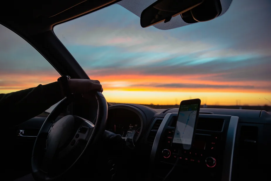 Una mano sujeta un volante de un vehículo en marcha mientras a través de la luna del coche se observa un atardecer.