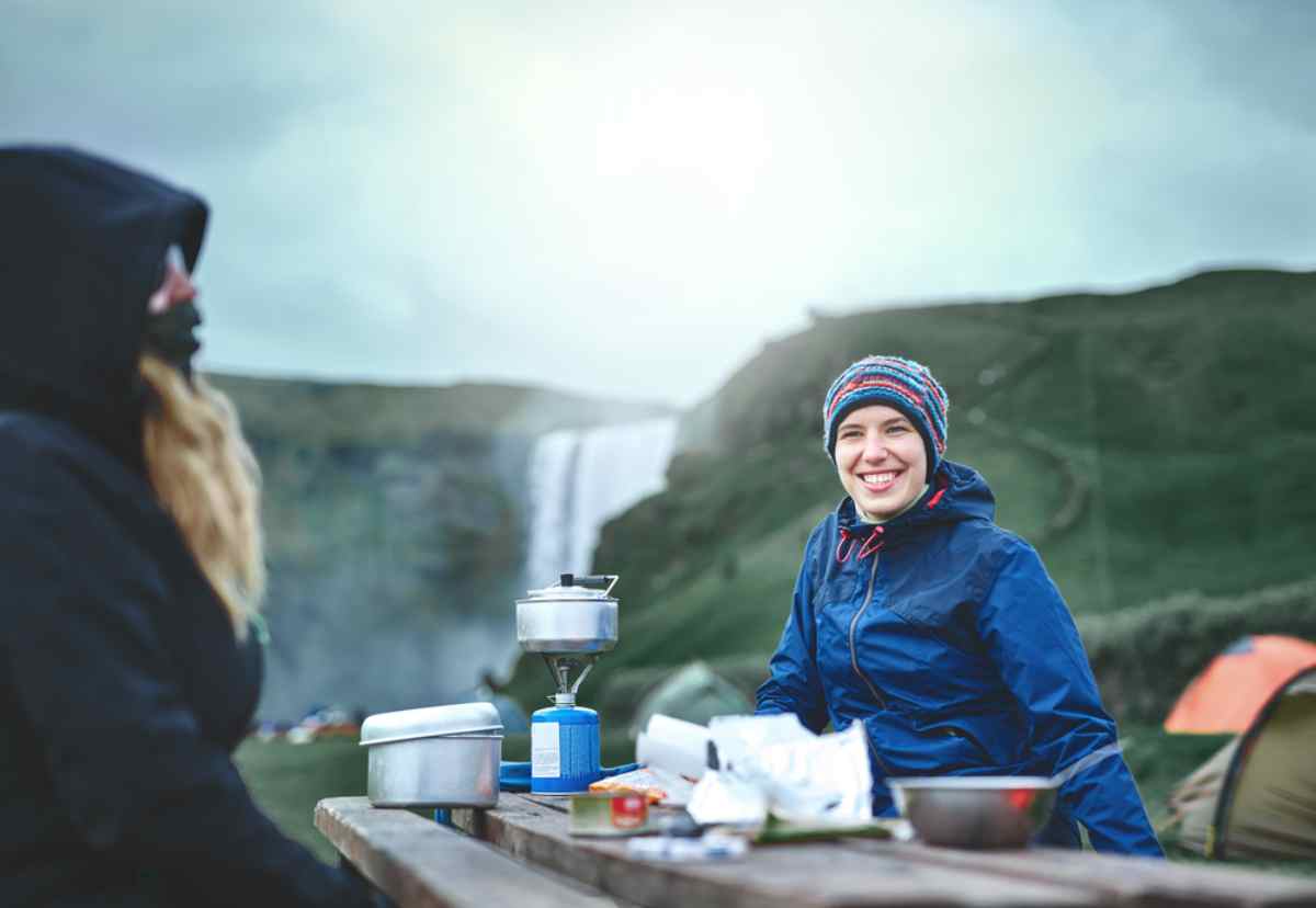 Cooking essentials for camping in Iceland Smiling camper at a picnic table cooking on a portable stove near a waterfall in Iceland, with cold weather layers and tents in the background.