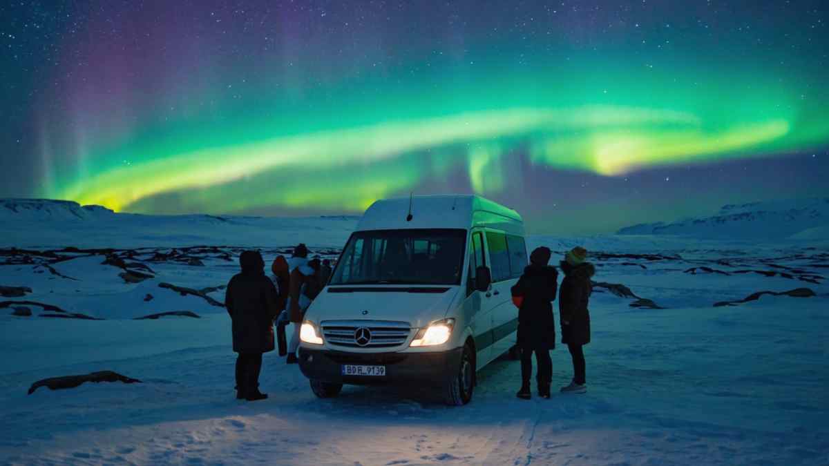 Camping in Iceland under northern lights White campervan on snowy plains while a small group watches the Northern Lights ripple across a starry Icelandic sky.