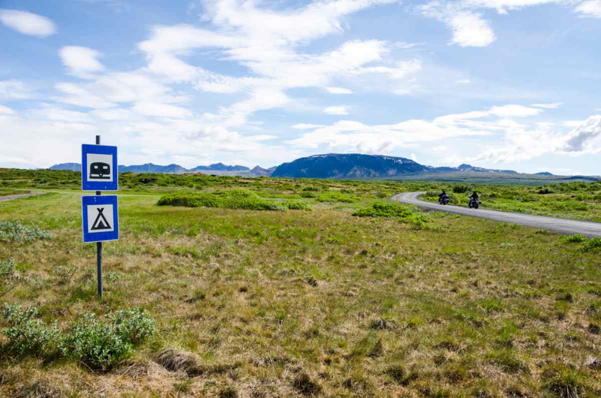 Camping sign in Iceland Blue roadside signs indicating motorhome and tent camping in rural Iceland, with motorcycles on a winding road and summer mountains ahead.