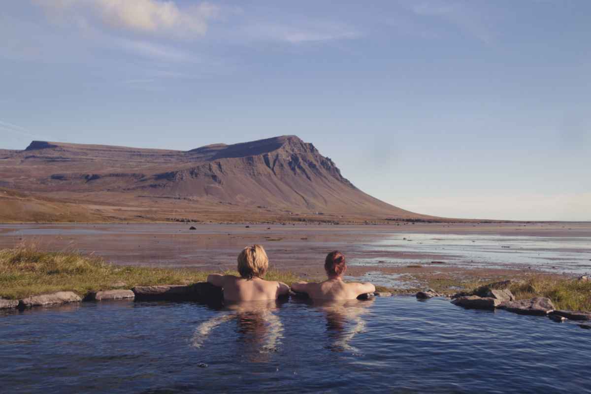 Two people relaxing in a natural hot spring with mountains and coastline ahead.