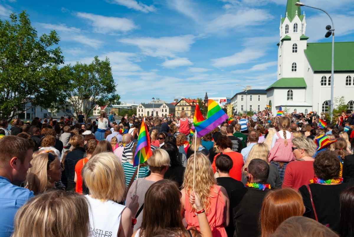 Crowd celebrating Reykjav&iacute;k Pride with rainbow flags near a white church.
