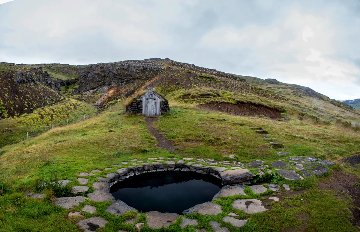 Gudrunarlaug hot spring in Iceland, featuring a small stone pool surrounded by green hills and a rustic wooden hut under a cloudy sky