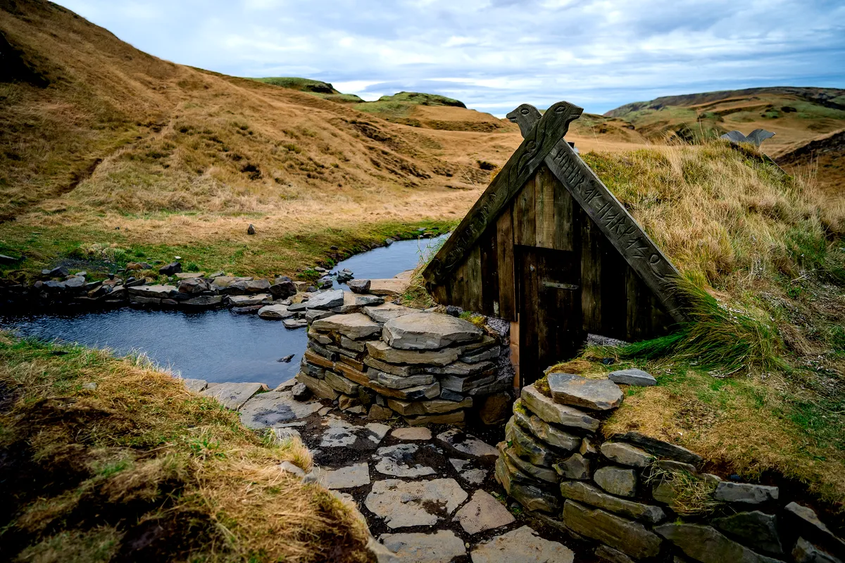 Hrunalaug hot spring in Iceland, featuring a rustic wooden hut beside a small stone-lined pool in a grassy landscape under a cloudy sky.