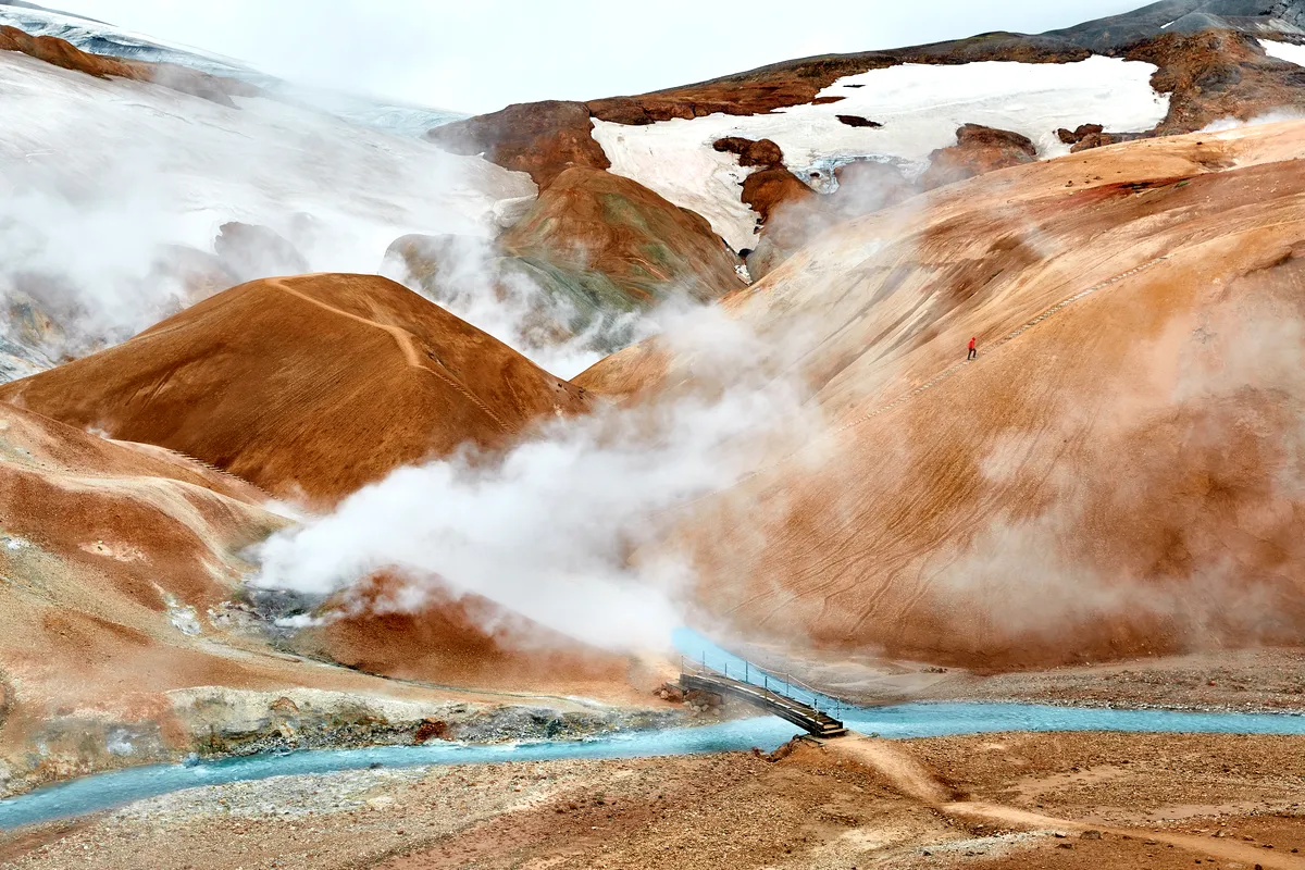 Steaming geothermal area of Landmannalaugar in Iceland, featuring colorful hills, a small wooden bridge over a blue stream, and patches of snow