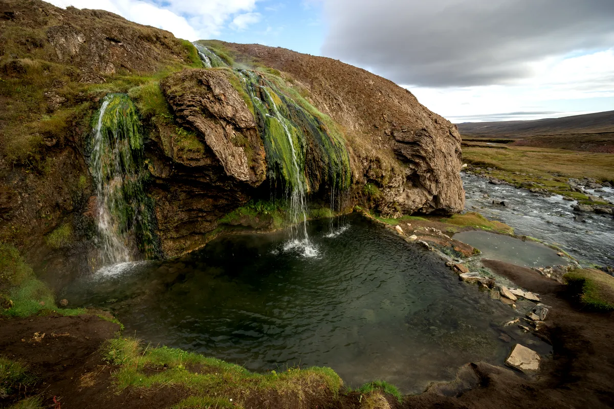 Laugavallalaug hot spring in Iceland, with water cascading over a moss-covered rock into a natural pool, set in a scenic landscape