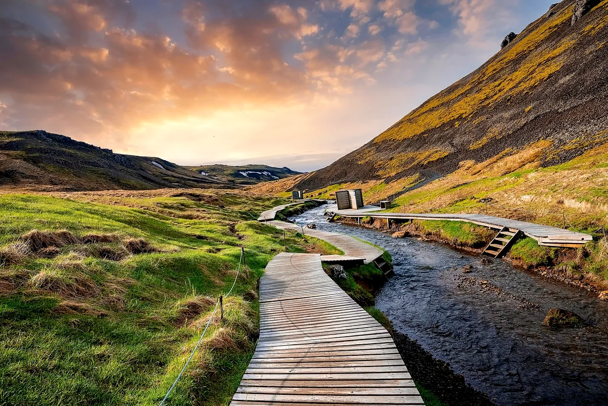 Wooden boardwalk along Reykjadalur River in Iceland, winding through a green valley with a beautiful sunset sky.