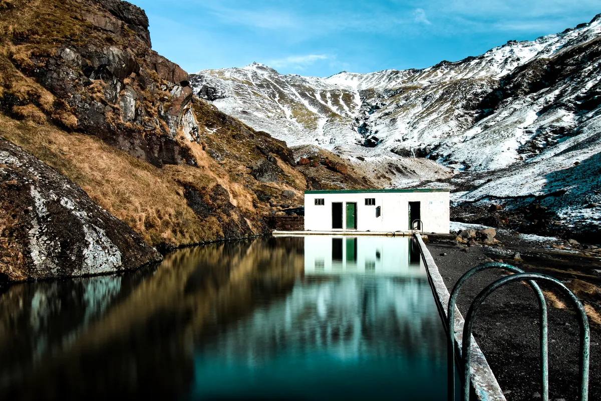 Seljavallalaug swimming pool in Iceland, nestled in a mountain valley with snow-capped peaks and a clear blue sky