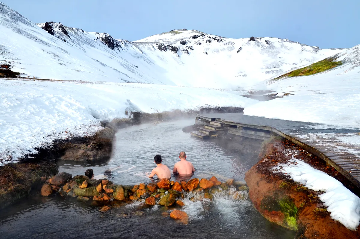 People bathing in the Reykjadalur hot springs in Iceland, surrounded by snowy mountains and a wooden boardwalk.