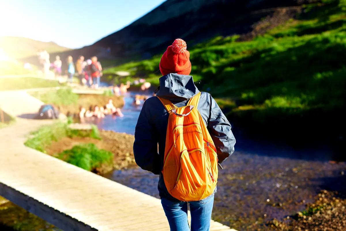 Person with a backpack and a red beanie hiking towards the Reykjadalur hot springs in Iceland, with people bathing in the background