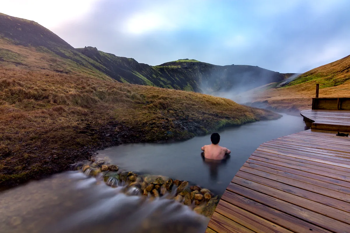 Person bathing in the Reykjadalur hot spring in Iceland, surrounded by mist and mountains, with a wooden boardwalk in the foreground.