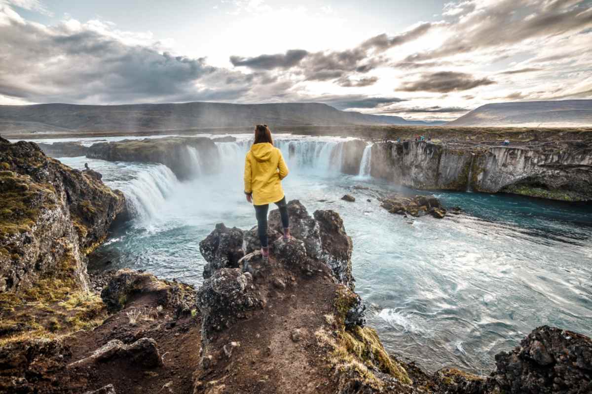 A woman in a yellow jacket stands motionless on a high rock in front of a spectacular waterfall in Iceland and a cloudy sky.