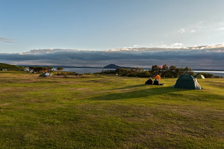 A scenic campsite in Iceland with tents set up on lush green grass, overlooking a peaceful lake and distant mountains—illustrating the beauty of Iceland’s designated campgrounds for campervan travelers.