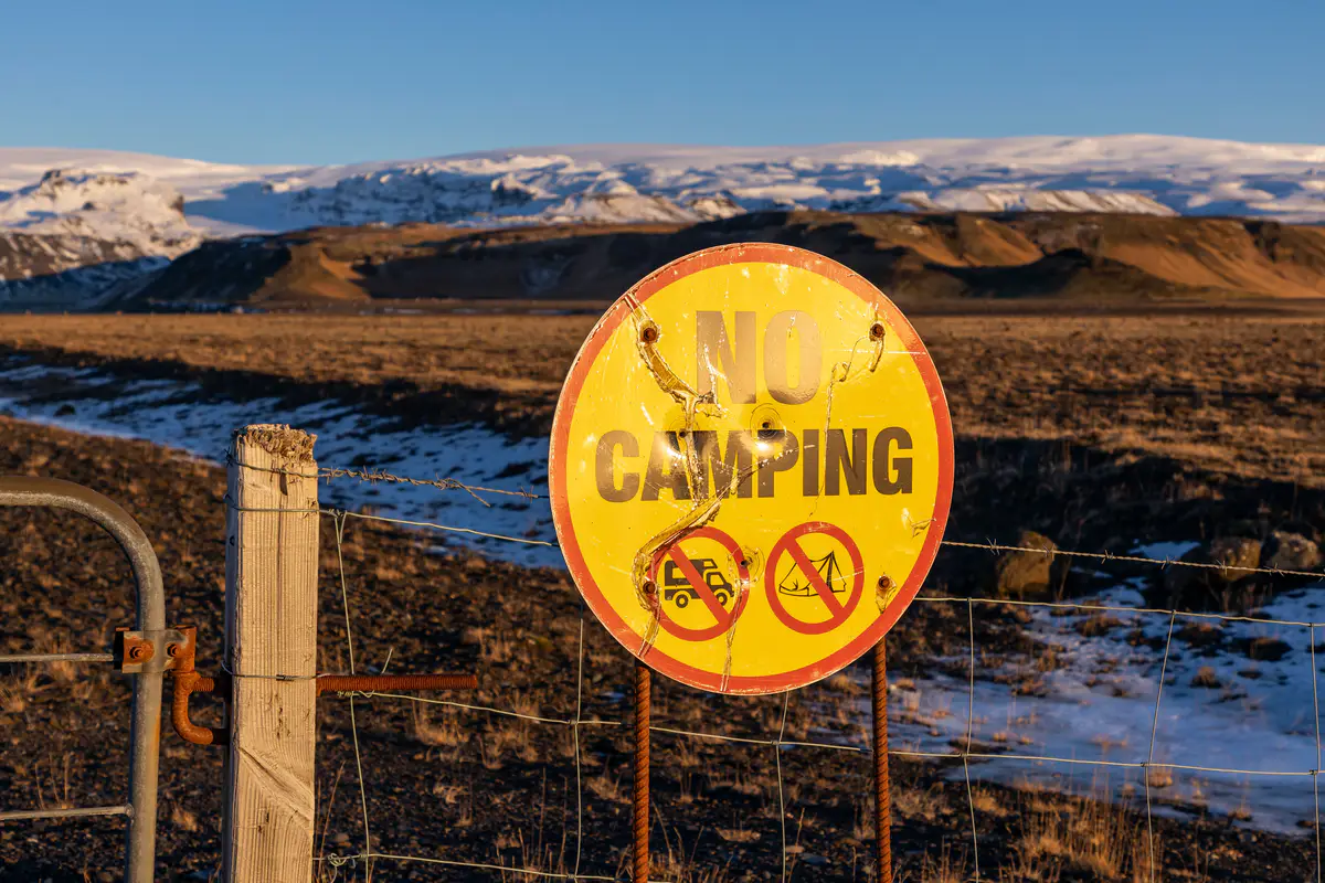 A weathered “No Camping” sign on a fenced-off area in Iceland, with snow-covered mountains in the background—highlighting the strict regulations against wild camping.