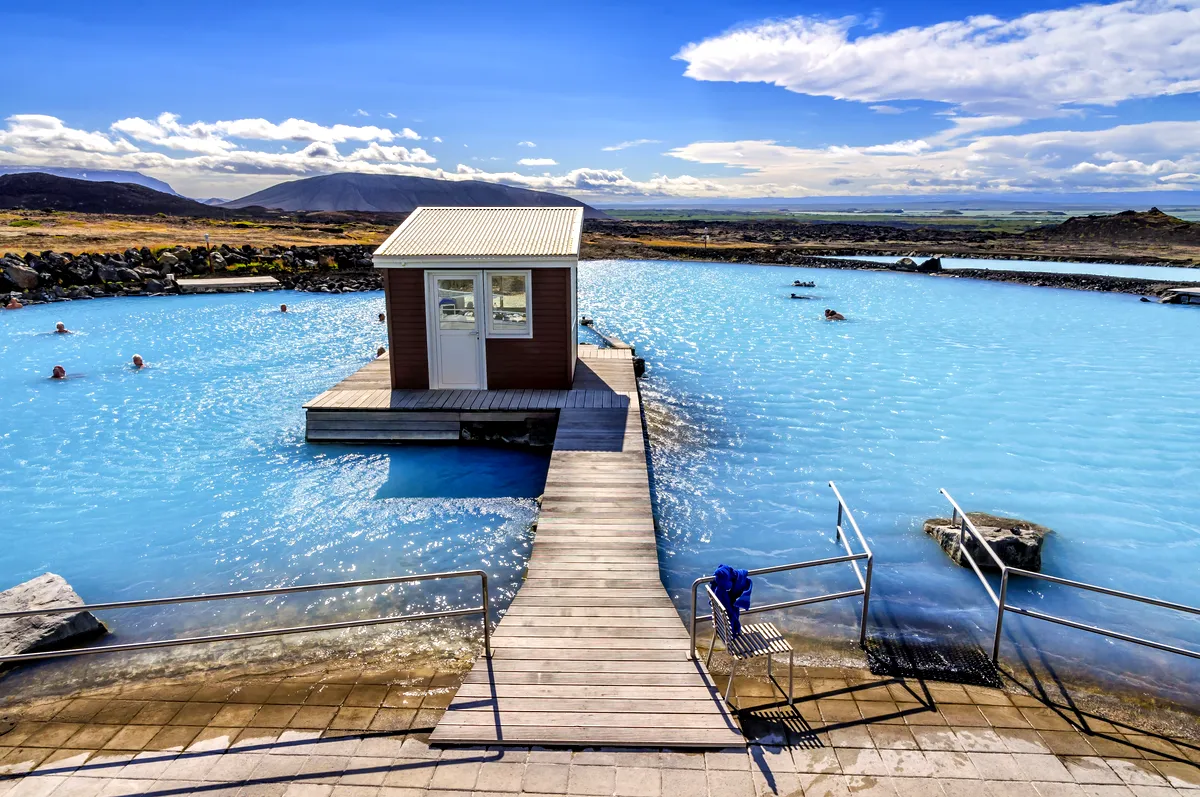 Laguna geotérmica de Myvatn bajo un cielo despejado, con aguas azules brillantes que reflejan la luz del sol. La imagen captura el pequeño edificio central rodeado de piscinas, con montañas y un vasto paisaje escénico de fondo