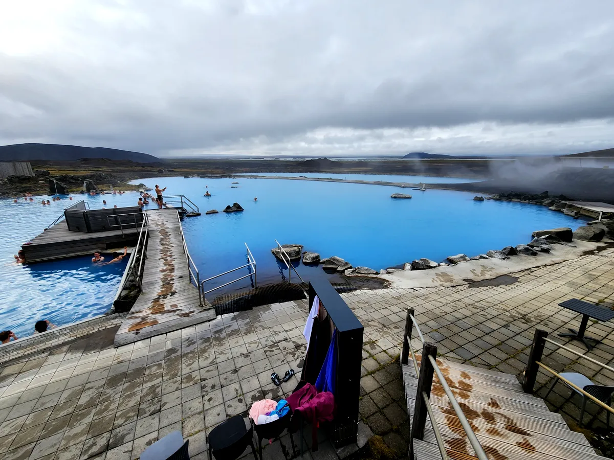 Vista panorámica del spa geotermal de Myvatn en Islandia, con sus enormes piscinas azules fundiéndose con el horizonte brumoso. Las plataformas de madera rodean las piscinas y ofrecen a los visitantes una experiencia envolvente y relajante. El paisaje circundante es accidentado, pero sereno, y ofrece un marcado contraste con las tranquilas aguas azules.