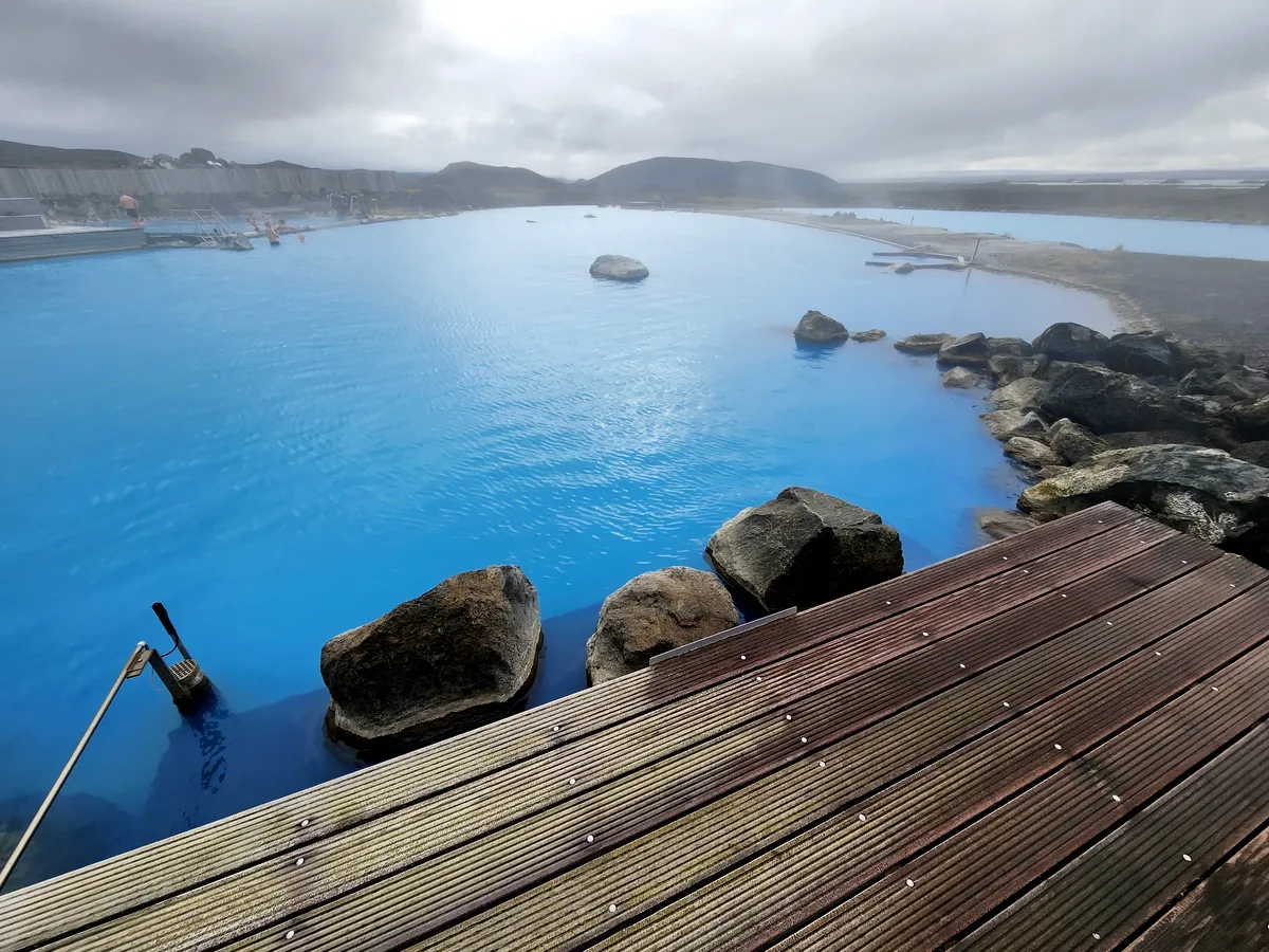 Una vista más cercana de las tranquilas aguas azules de la laguna geotérmica de Myvatn en Islandia. Las rocas y las pasarelas de madera crean una atmósfera tranquila mientras el vapor se eleva desde las cálidas aguas, mientras que el paisaje lejano, envuelto en niebla, añade un toque misterioso al entorno.