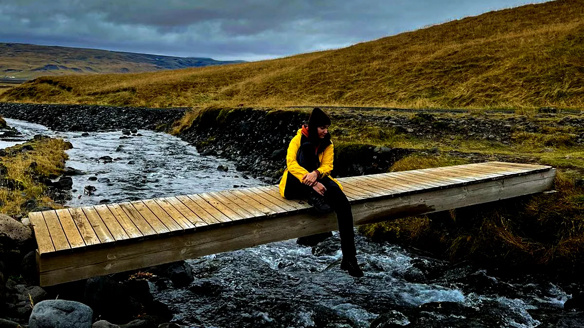 A traveler sits on a wooden bridge over a calm river in the Icelandic countryside