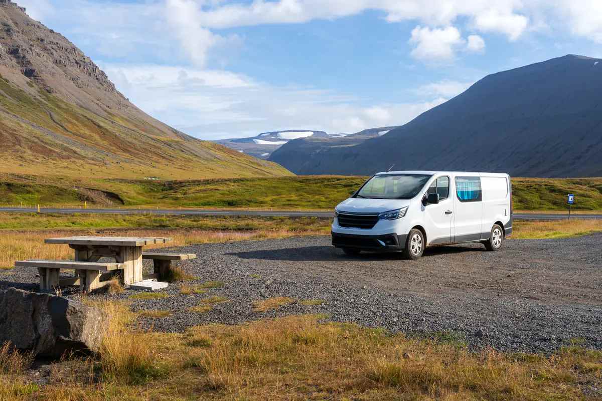 Una campervan blanca estacionada en un área de descanso designada en Islandia, con montañas y un glaciar al fondo. Cumpliendo con las leyes de camping en Islandia, el vehículo está estacionado en un lugar autorizado para evitar multas y daños ambientales.