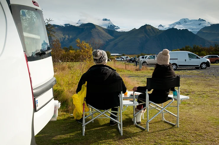 Dos viajeros disfrutando de un camping escénico en Islandia, sentados fuera de su campervan con vistas a montañas y glaciares. Un ejemplo perfecto de un camping bien equipado para viajeros en campervan.