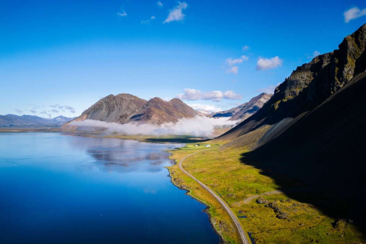 Scenic coastal road winding along calm blue water in Iceland&rsquo;s Eastfjords, framed by rugged mountains and low cloud.