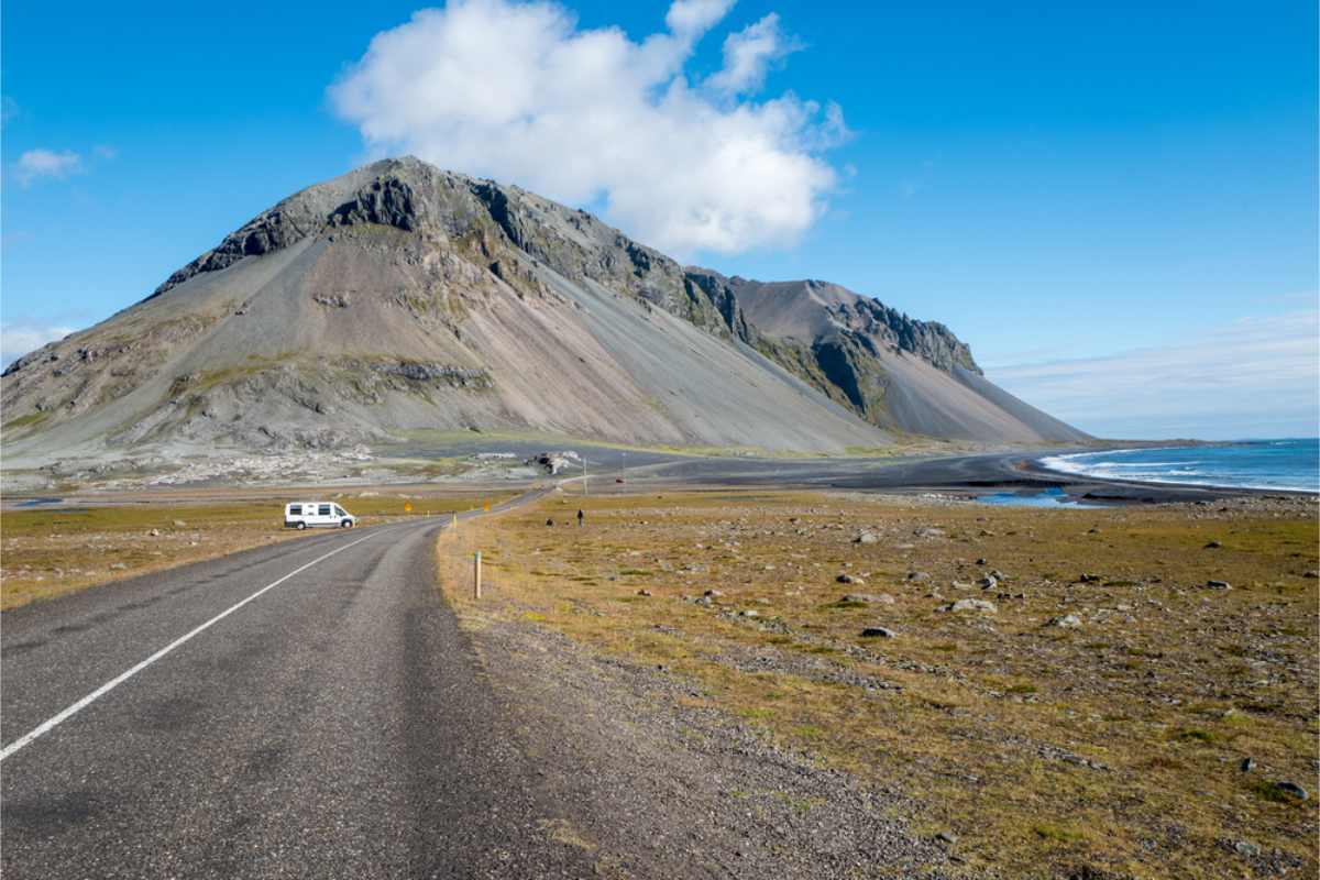 Open Ring Road stretching toward a steep mountain beside Iceland&rsquo;s coastline, with a small vehicle in the distance under a blue sky.