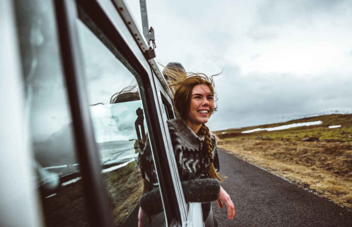 A red-haired woman pokes her head out of the window of a moving vehicle as the Icelandic wind blows her long hair.
