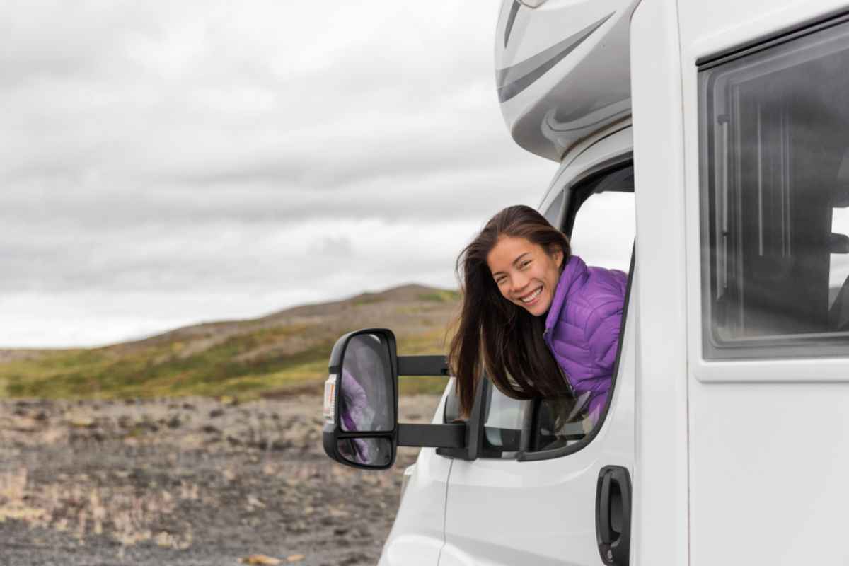 Une femme brune aux cheveux longs sort la tête par la fenêtre d'un camping-car en souriant à la caméra.