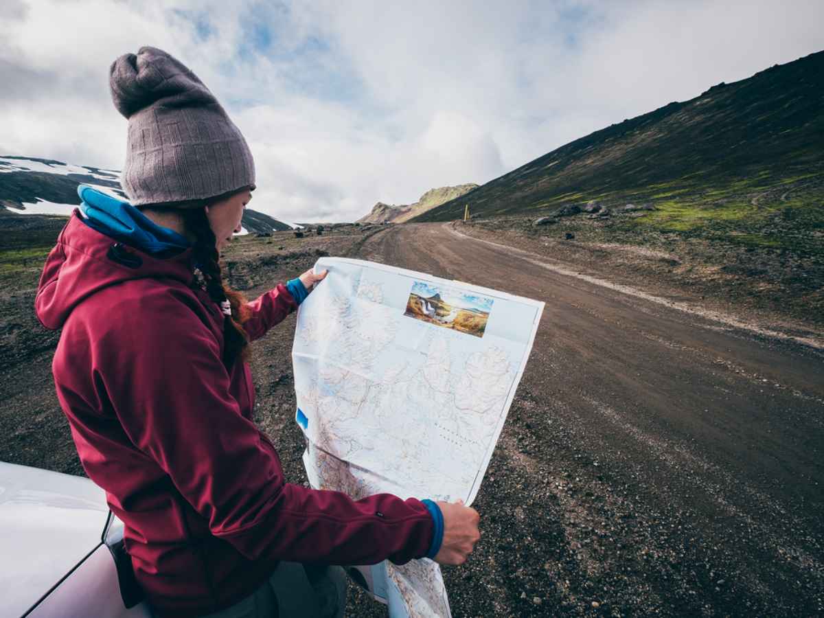 Une femme portant un bonnet et des tresses regarde une carte de l'Islande tandis que son véhicule est garé sur le bord de la route.