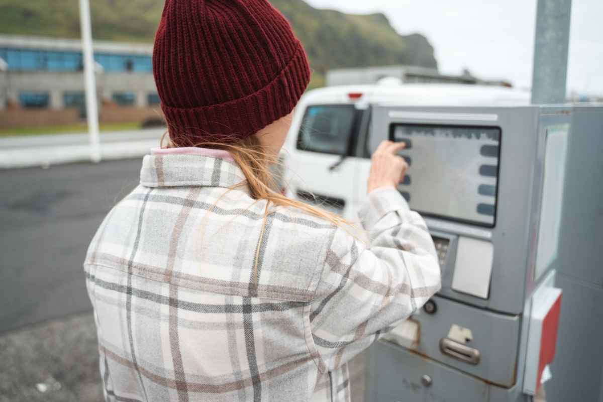A woman in a woolen cap with her back turned manipulating a machine at a petrol station in Iceland at what looks like the moment of paying for the refueling.