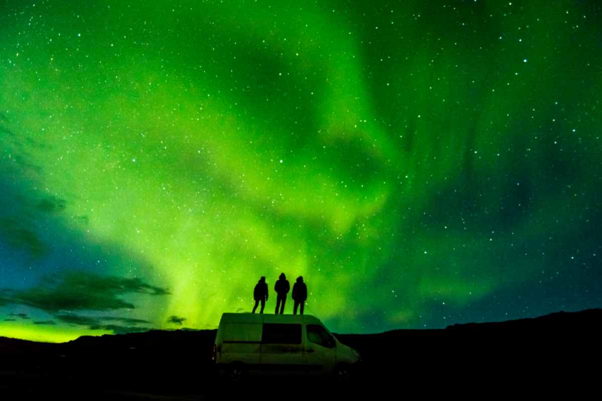 Tourists standing on top of a campervan watching green northern lights dance on Iceland's sky