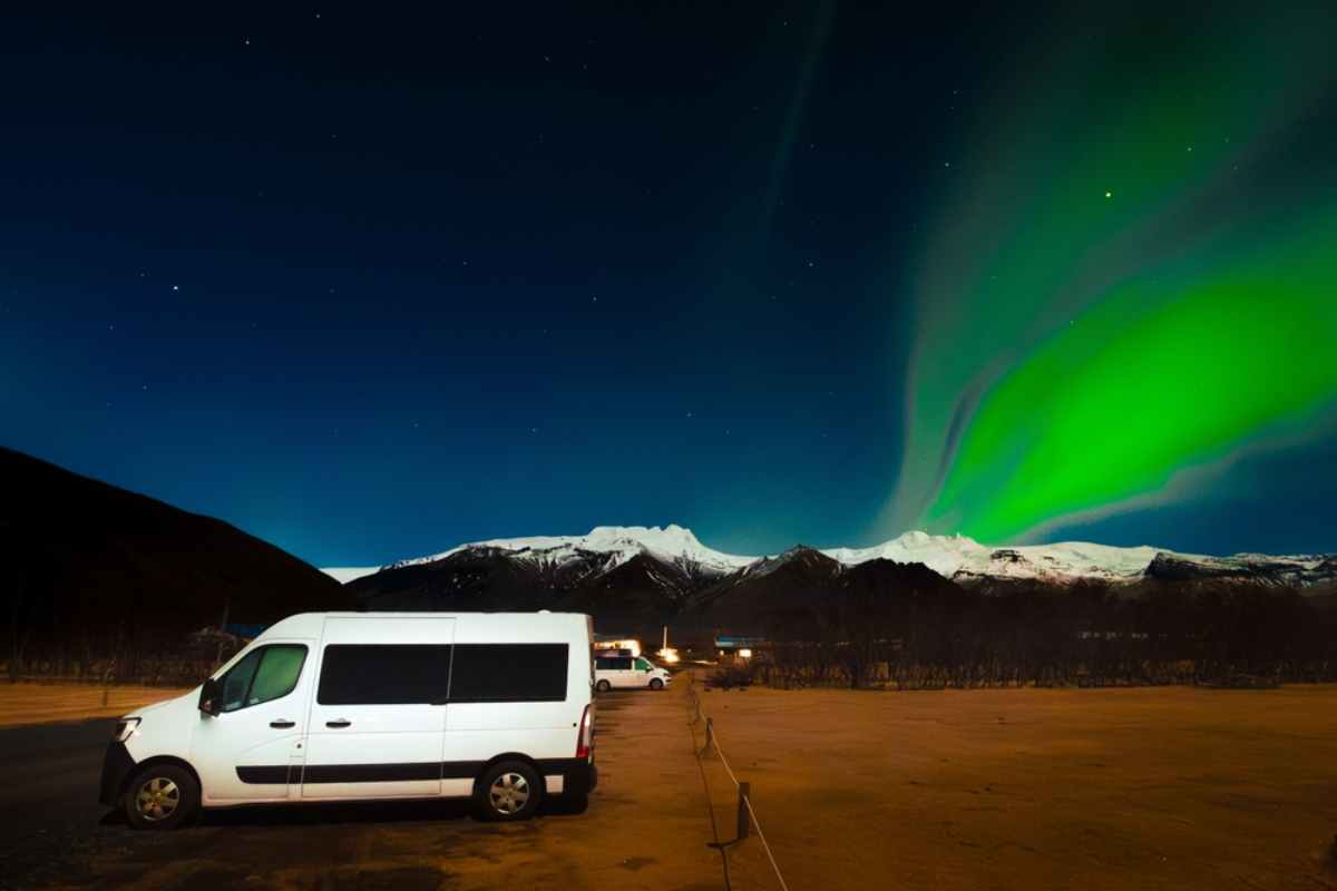 White camper parked by a mountain range with snow covered peaks and the Northern Lights dancing above