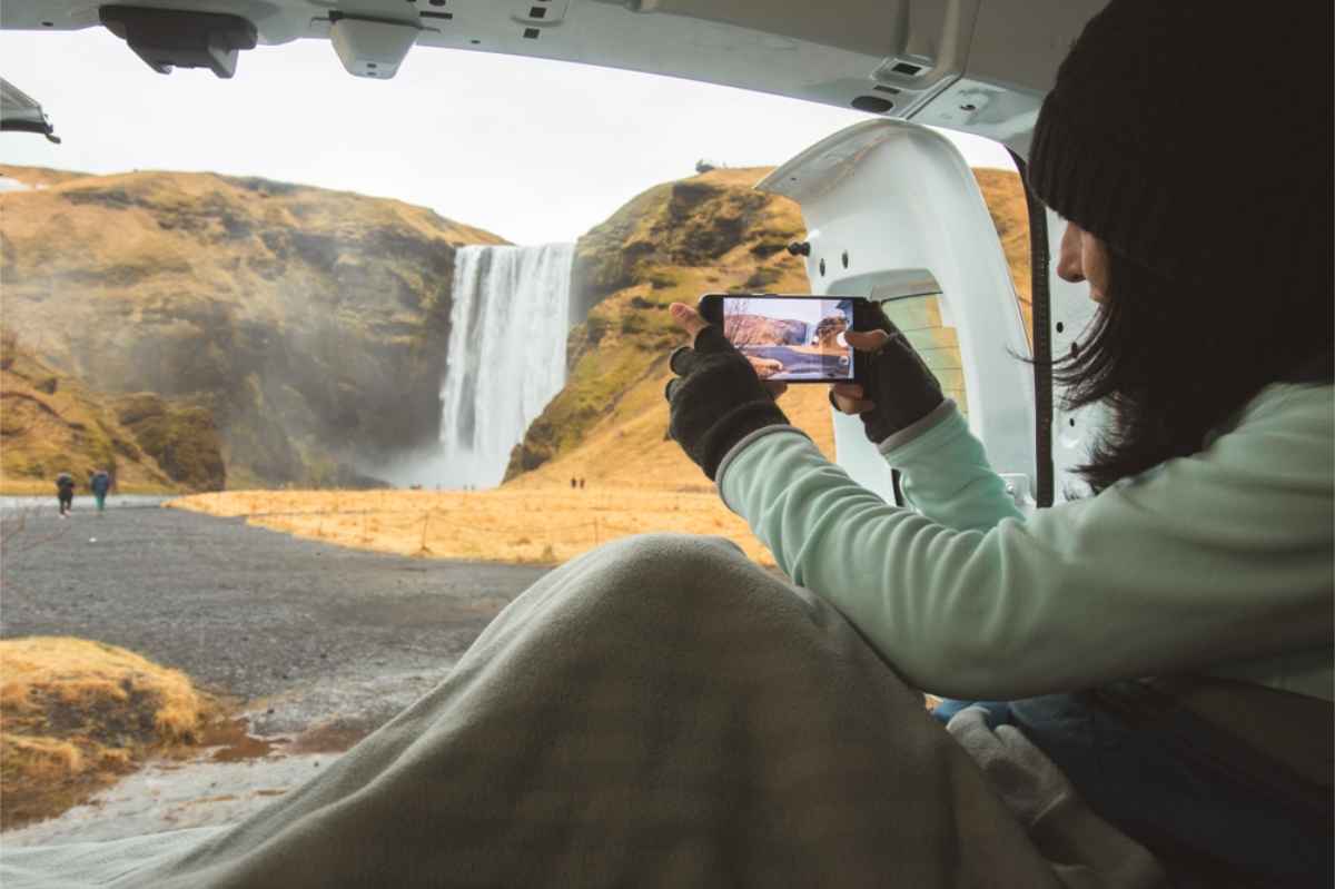 Girl sitting inside her campervan matress with the rear doors open while taking a picture of Skogafoss waterfall in Iceland