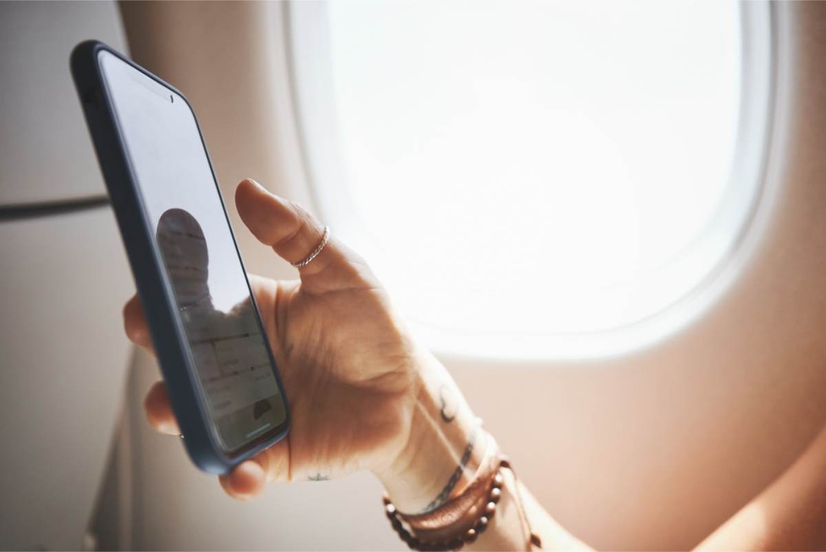 Checking a travel app on the plane Close-up of a hand holding a smartphone beside a bright aeroplane window, with stacked bracelets on the wrist