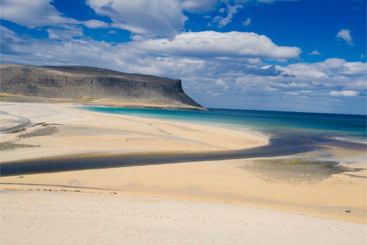 Golden sand beach below a flat-topped cliff Wide beach of pale sand with turquoise shallows and tidal channels, beneath a dark table mountain under scattered cloud