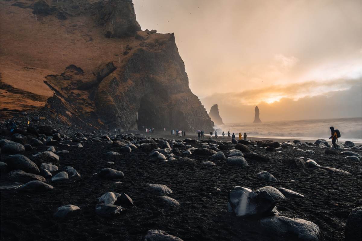 Black sand beach with sea stacks at sunset Visitors walking on black volcanic sand beside a basalt cliff, with tall rock stacks in the sea and low sun through the spray