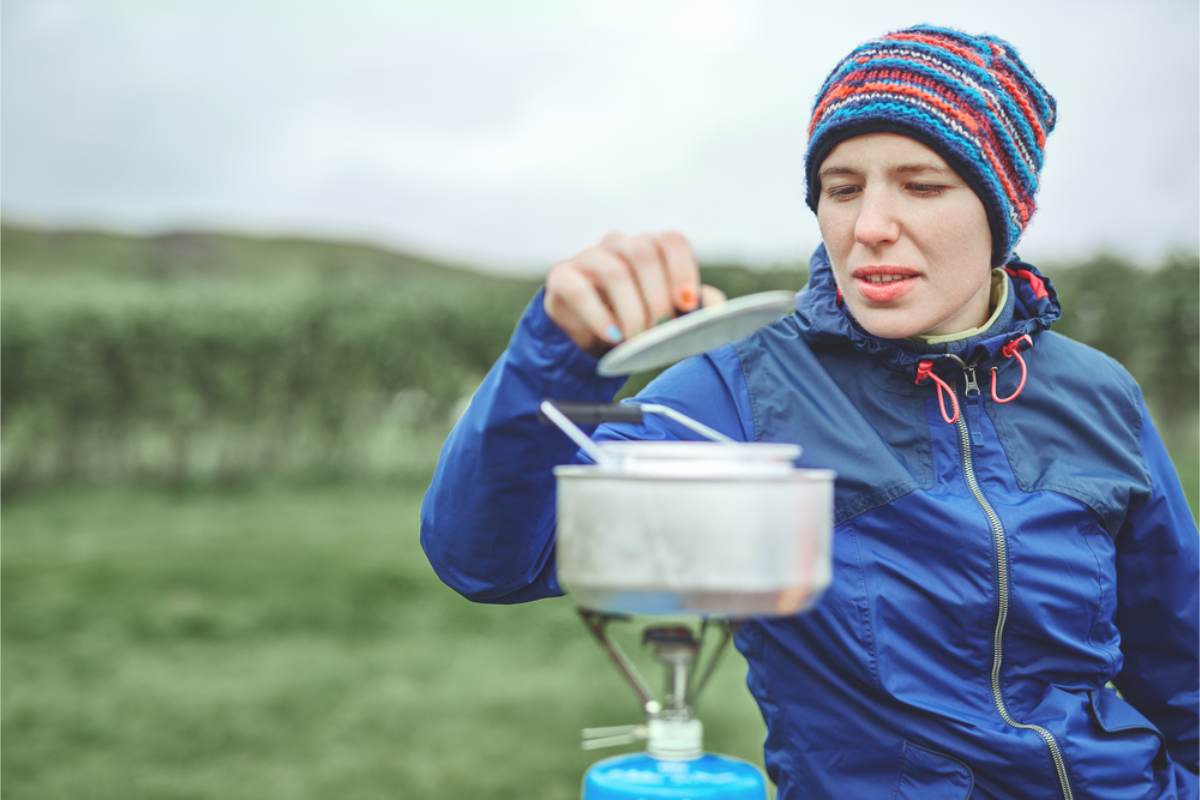 Cooking on a camping stove in Iceland Woman in a blue jacket and striped hat lifting the lid off a pot on a small gas camping stove outdoors