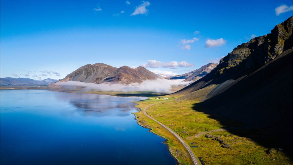 Coastal road along an Icelandic fjord Aerial view of a narrow road following a green shoreline beside still blue water, with low cloud around brown mountains