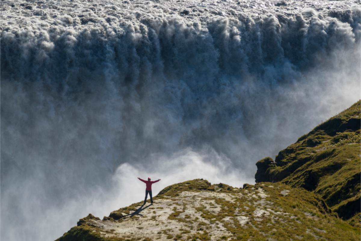 Standing beside a powerful Icelandic waterfall Person in a red jacket standing with arms outstretched on a green ridge in front of a wide, thundering waterfall and spray