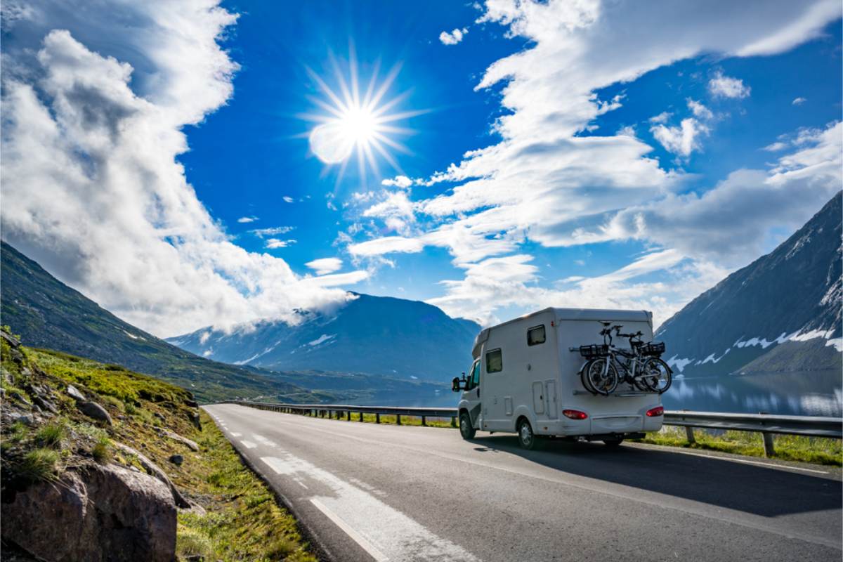 Motorhome on a mountain road in summer White motorhome with bikes on the back driving a road between mountains and a lake under a bright sun and blue sky