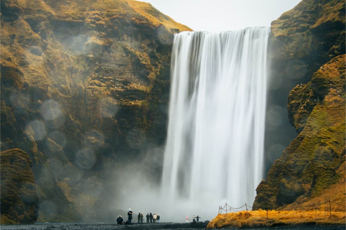 Waterfall on Iceland's south coast Tall waterfall dropping over a mossy cliff into a black sand basin, with a small group of visitors at the base for scale