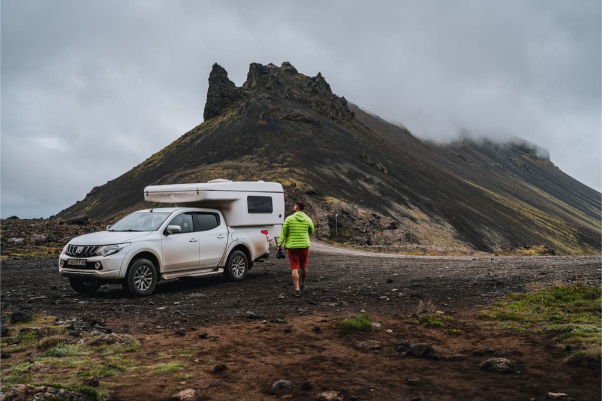 Pickup truck camper below a volcanic peak Man in a green jacket walking toward a white pickup with a truck camper, parked on gravel at the foot of a dark ridge