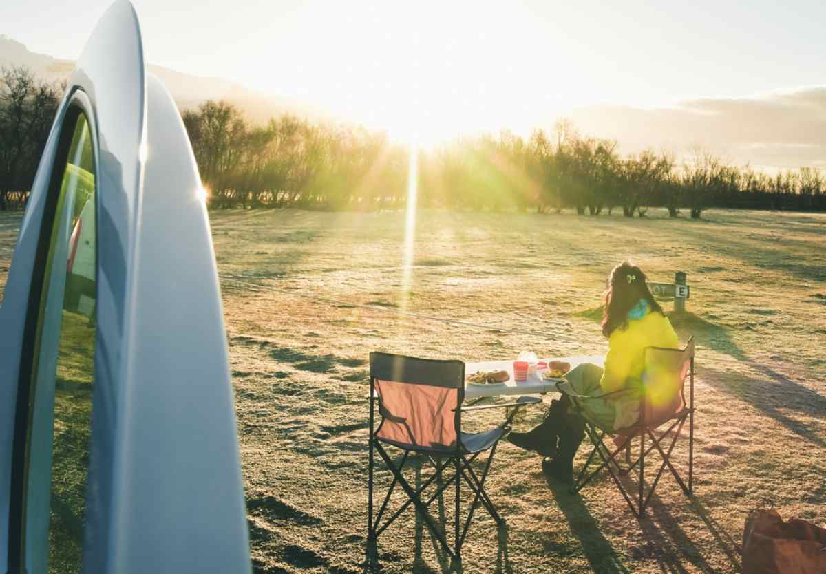 Woman in a yellow jacket eating breakfast at a folding table beside a camper at a frosty campground at sunrise.