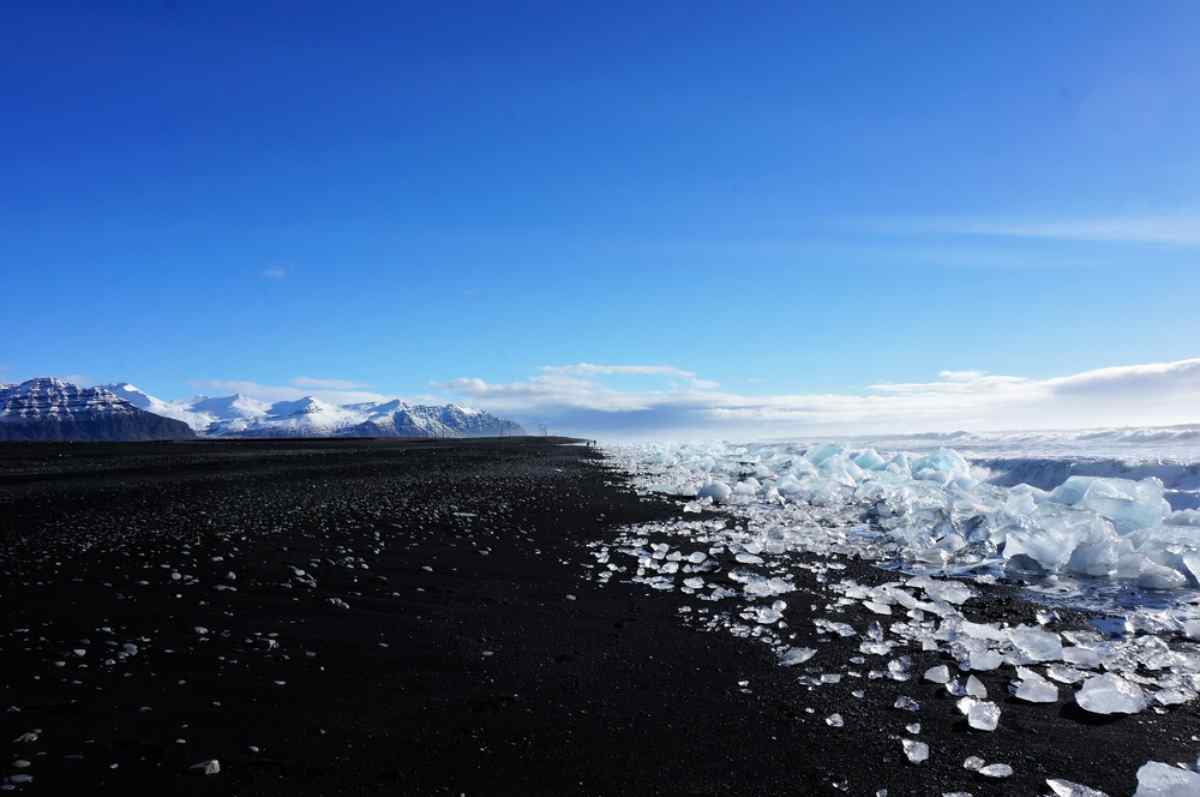 Glacial ice chunks scattered across black sand with snowy mountains along Iceland’s coast.