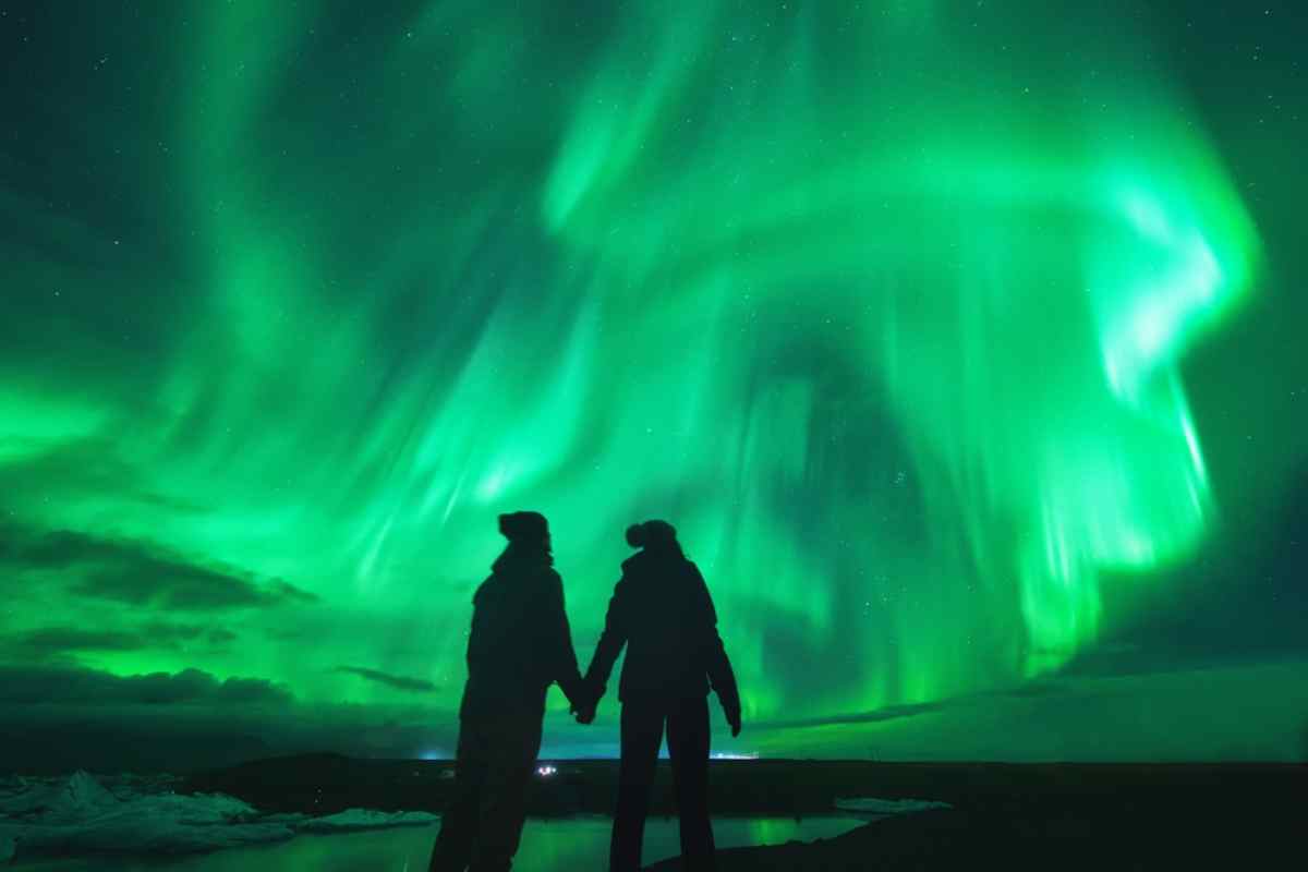 Silhouette of a couple holding hands while bright green aurora borealis swirls overhead and reflects on water.