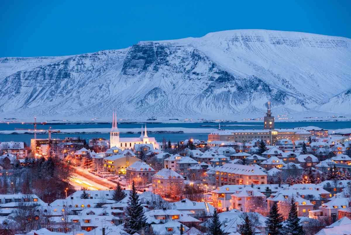 Blue-hour view of Reykjavik blanketed in snow, warm city lights and church spires with Mount Esja across the bay.