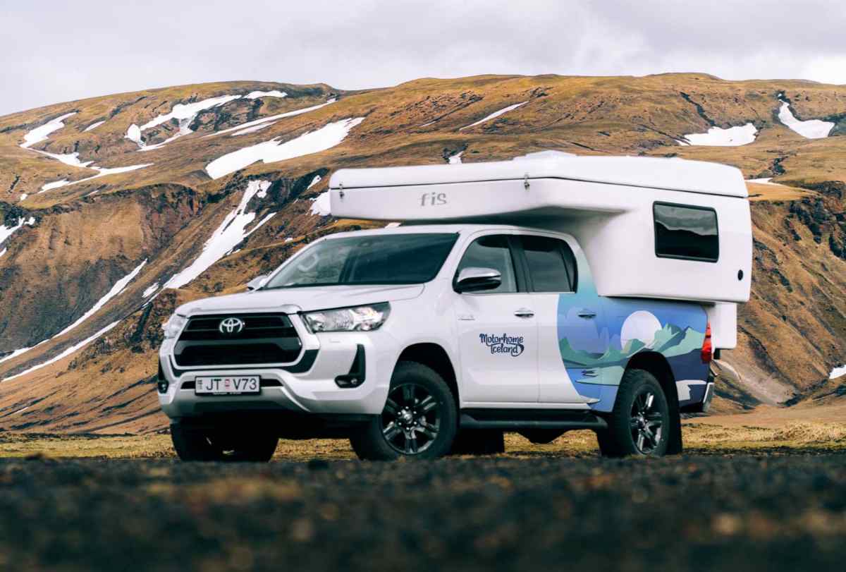 White Toyota Hilux with a slide-in camper parked on dark gravel, rugged Icelandic mountains with patchy snow in the background.