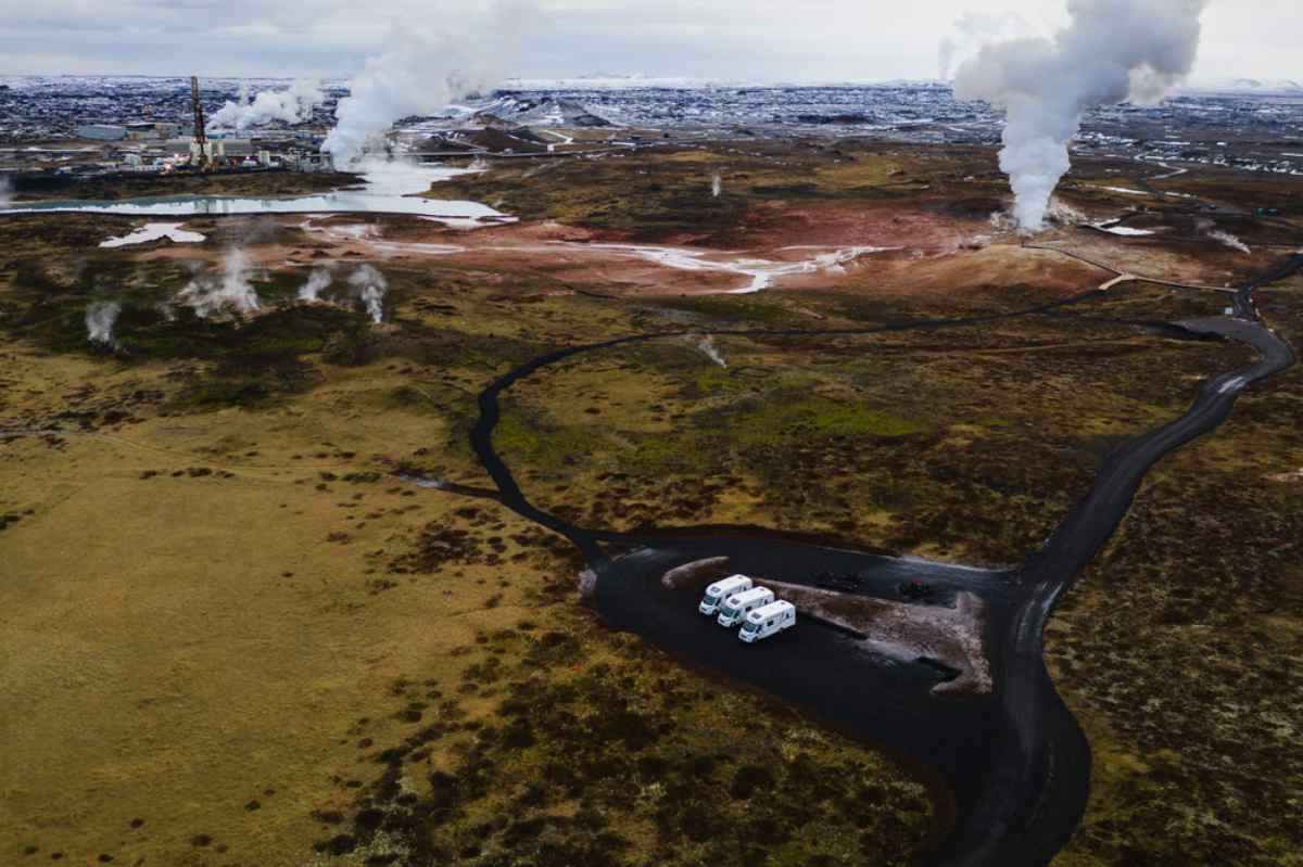 Aerial of steaming geothermal field with four white motorhomes parked on a black gravel turnout and a winding winter road.