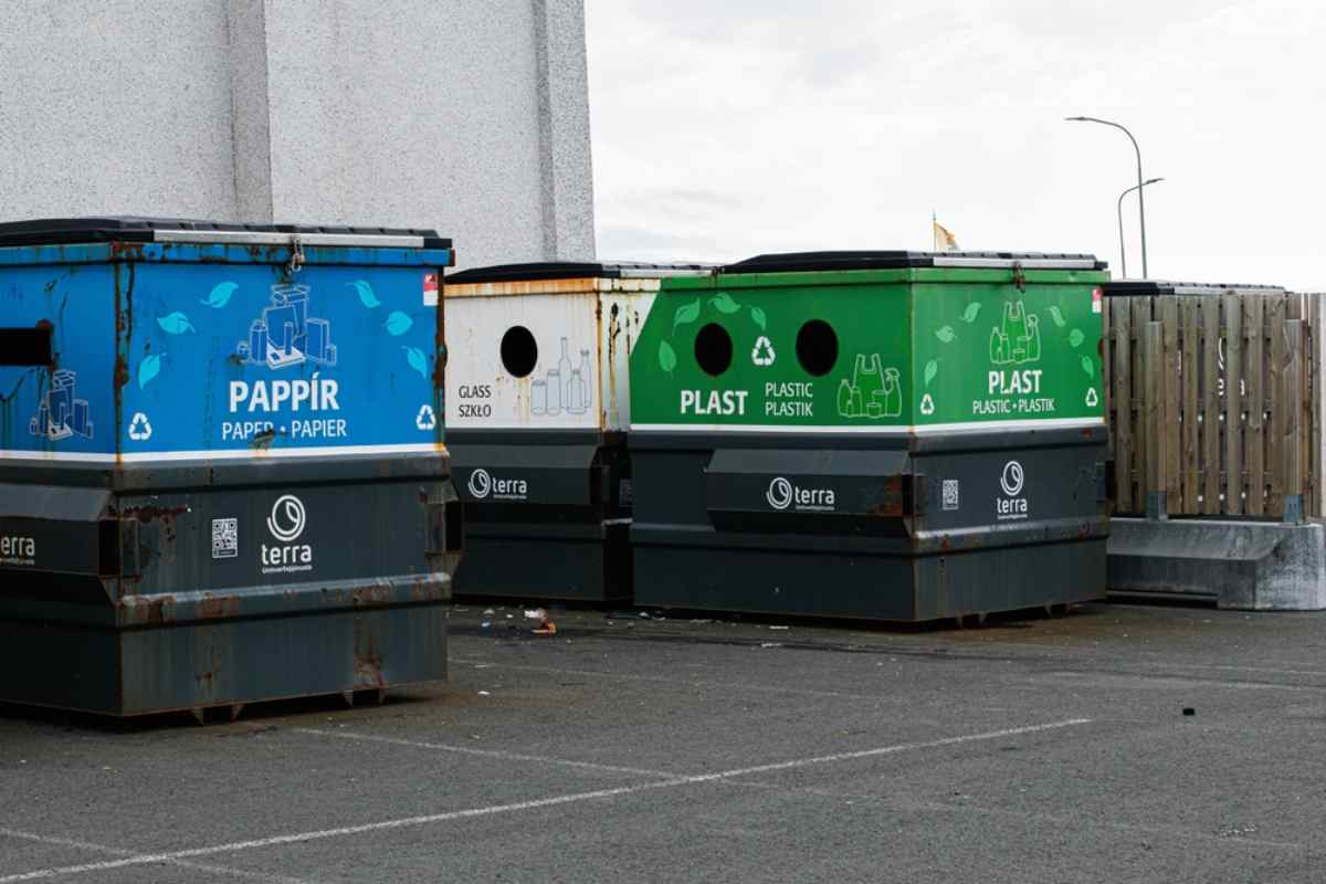 Color-coded containers for paper, glass, and plastic recycling in an Iceland parking area.